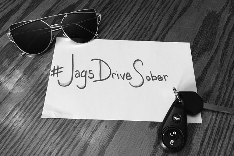 Sign with the hashtag "Jags Drive Sober" on a table with sunglasses and keys.