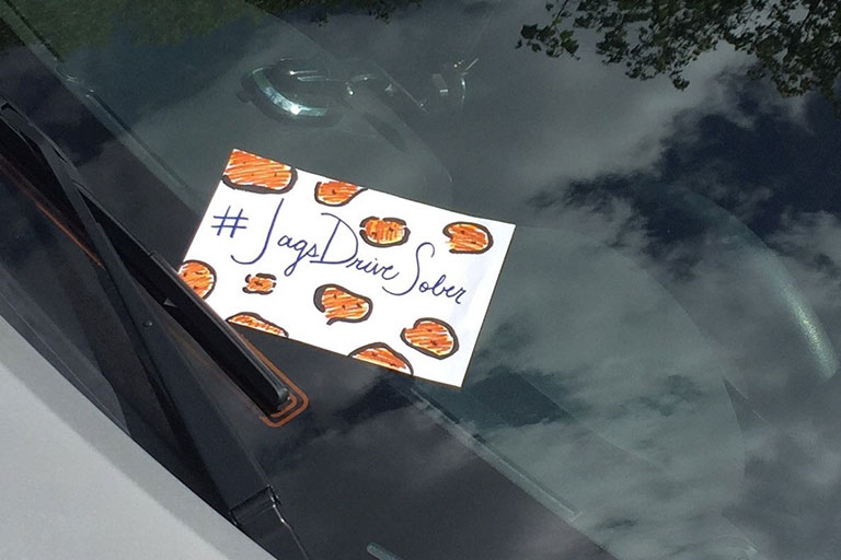 Postcard sized sign with the with the hashtag "Jags Drive Sober" on a windshield of a car.